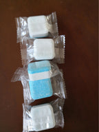 Four individually wrapped dishwasher tablets on a wooden surface, mostly white with one blue tablet