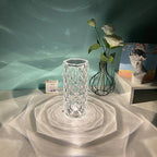 Clear glass textured vase casting light patterns on white surface, with wire-frame vase holding white flowers, decorative bust, and small calendar in background
