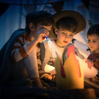 Three children reading by flashlight inside a cozy indoor tent with paper flames